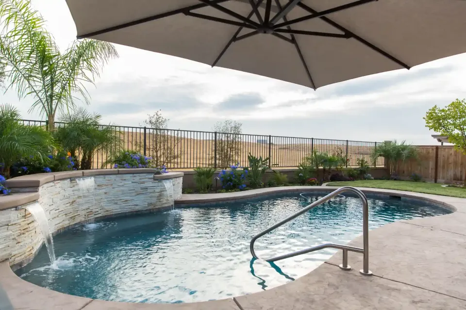 Outdoor pool with a curved design, umbrella shade, and patio seating beside a fenced view of the landscape
