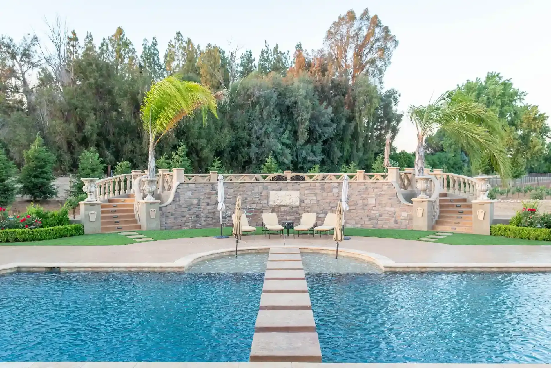 Stone walkway across a blue pool leading to a landscaped garden with palm trees and a stone wall