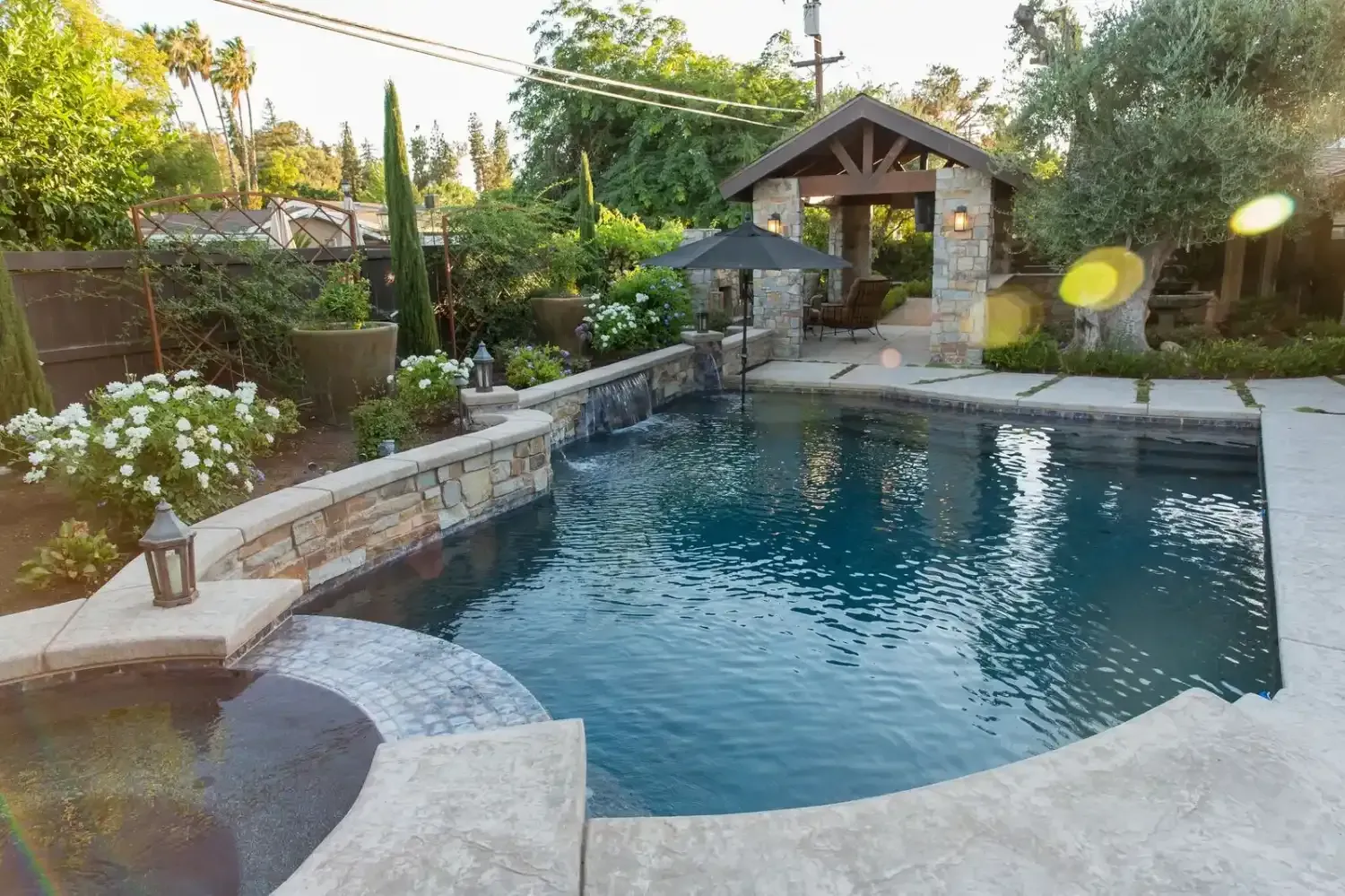 Backyard swimming pool with stone patio, small spa, and garden pergola at dusk