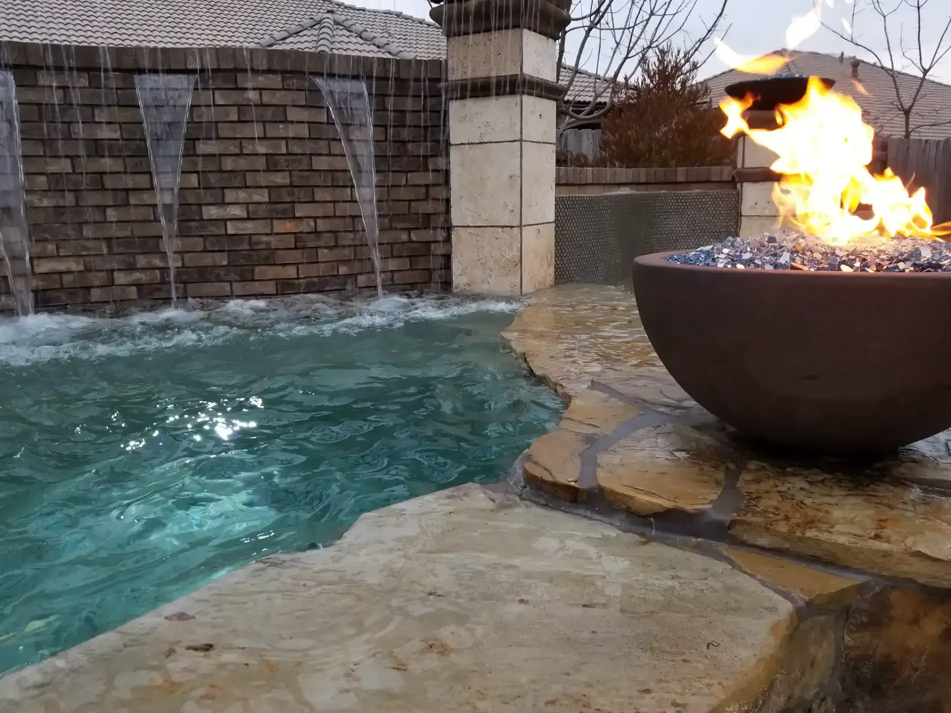 Stone pool with turquoise water beside a fire bowl on a patio, with a brick wall and waterfall feature.
