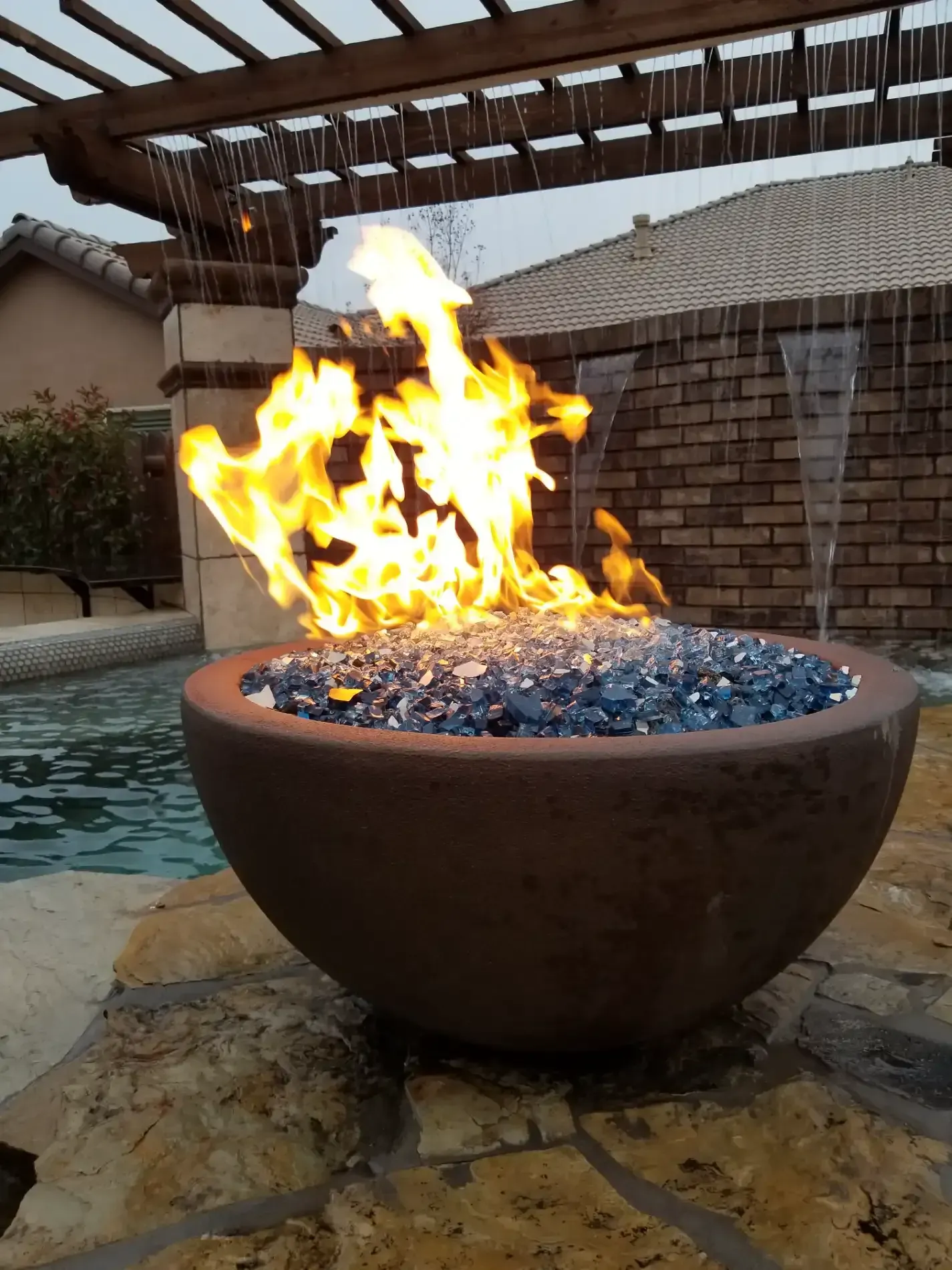 Flames rising from a round fire bowl beside a pool under a pergola at dusk