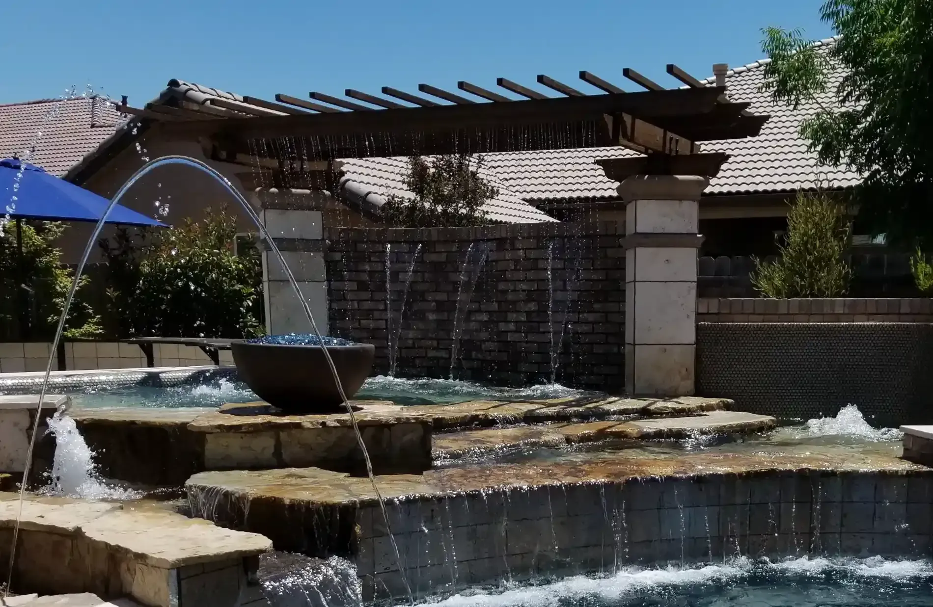 Outdoor fountain with cascading water, stone tiers, and a pergola in a sunny courtyard