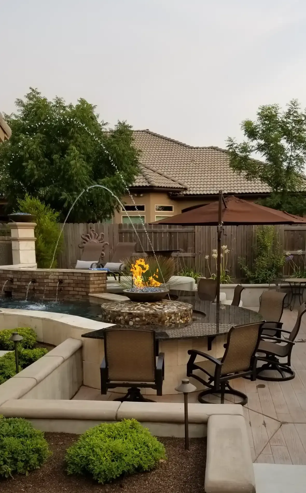 Backyard patio with beige seating, fire pit, and pergola near a house and trees.