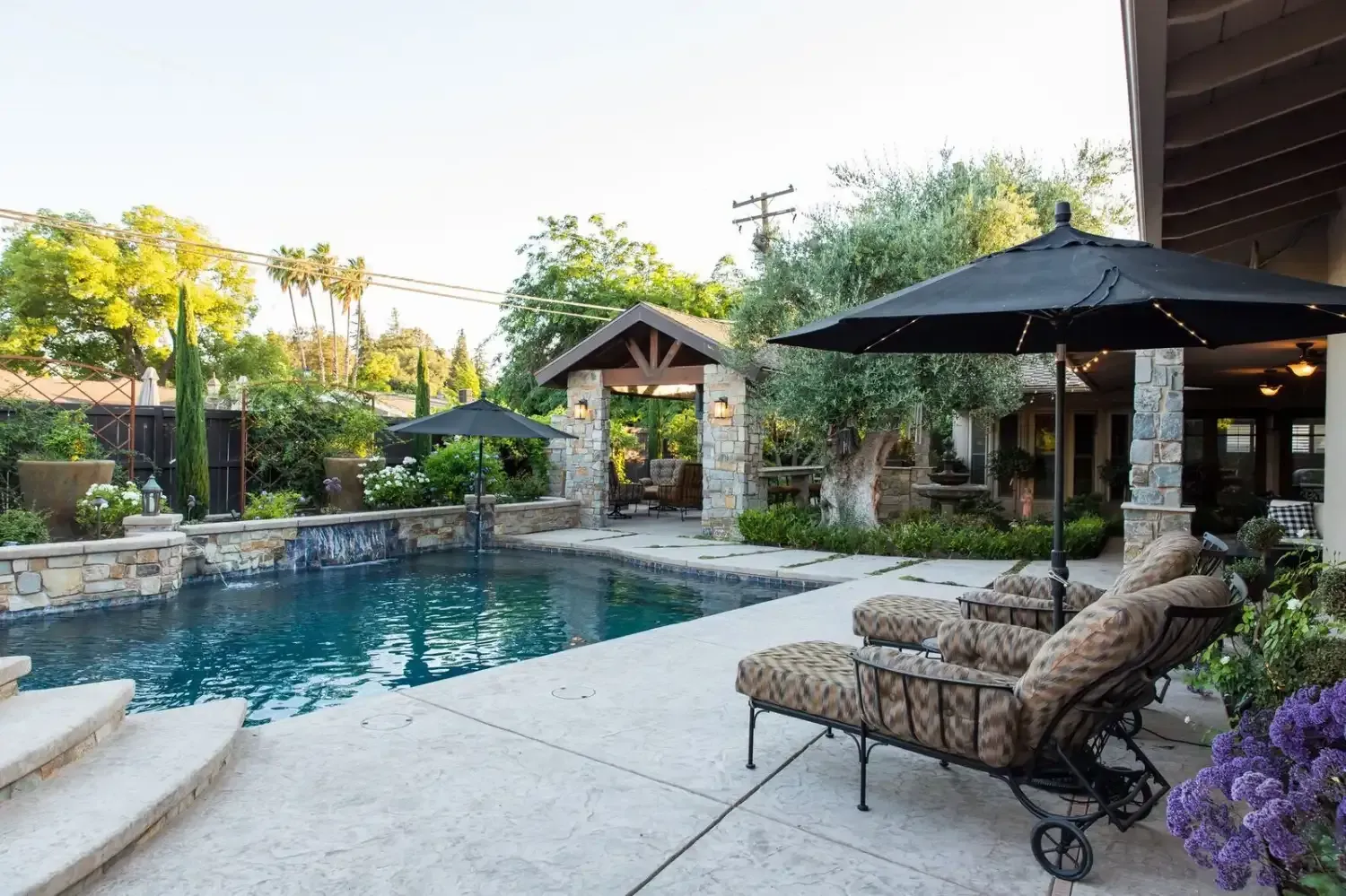 Backyard pool with stone patio, lounge chairs, umbrellas, and lush garden greenery at sunset