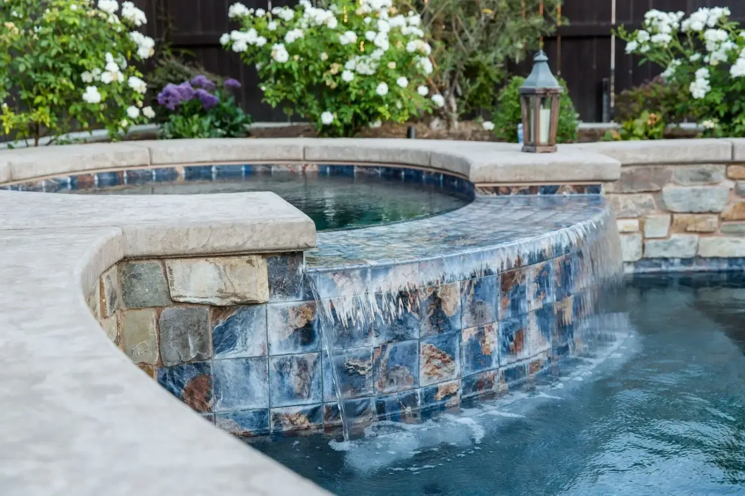 Backyard stone waterfall spilling into a blue pool with lush greenery and lanterns