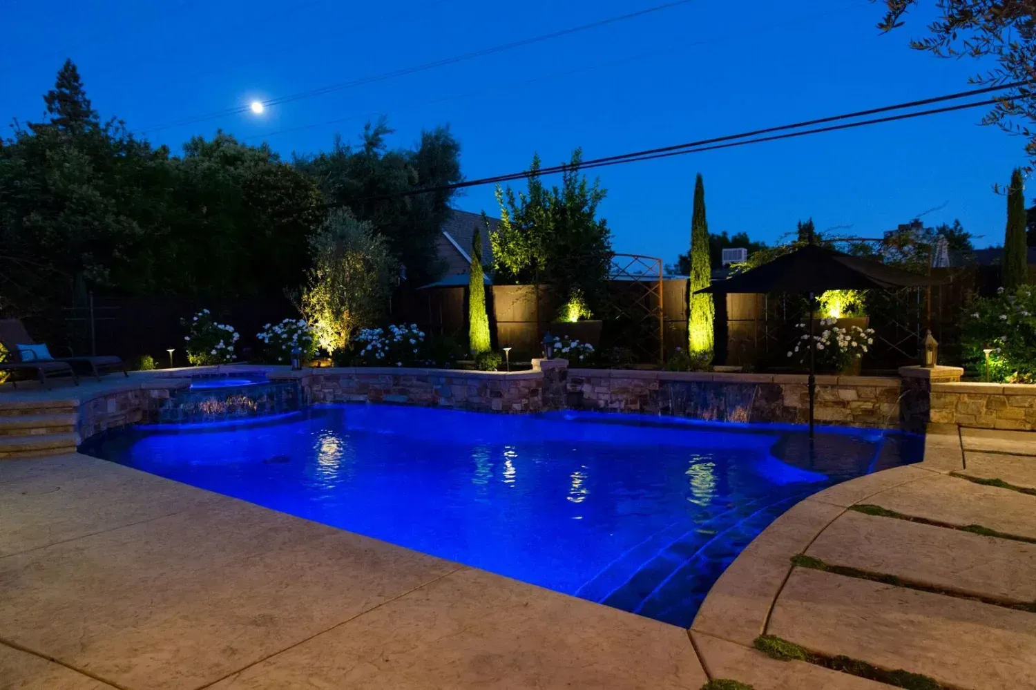 Backyard pool glowing blue at night, with stone patio and lit landscaping around it