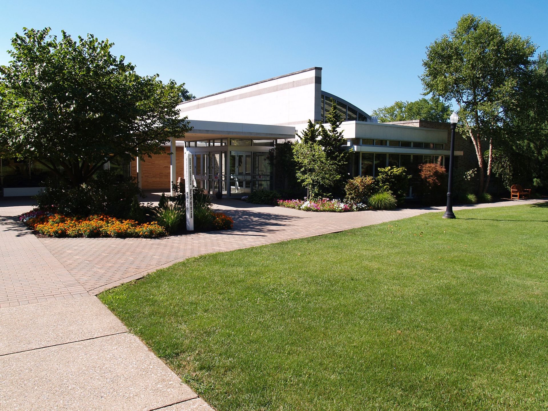 Modern building entrance with white canopy, landscaped flower beds, and a curved walkway beside green lawn