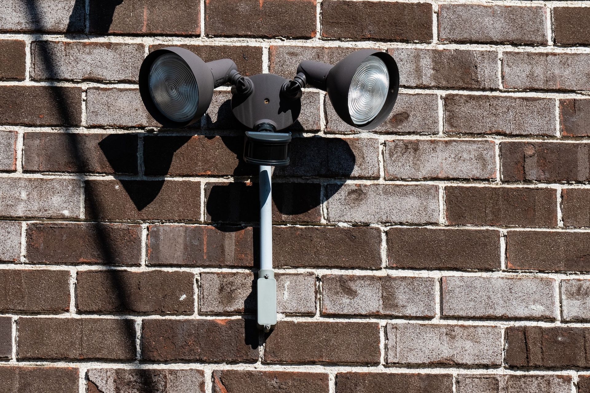 Black twin floodlight mounted on a brick wall, with a central motion sensor.
