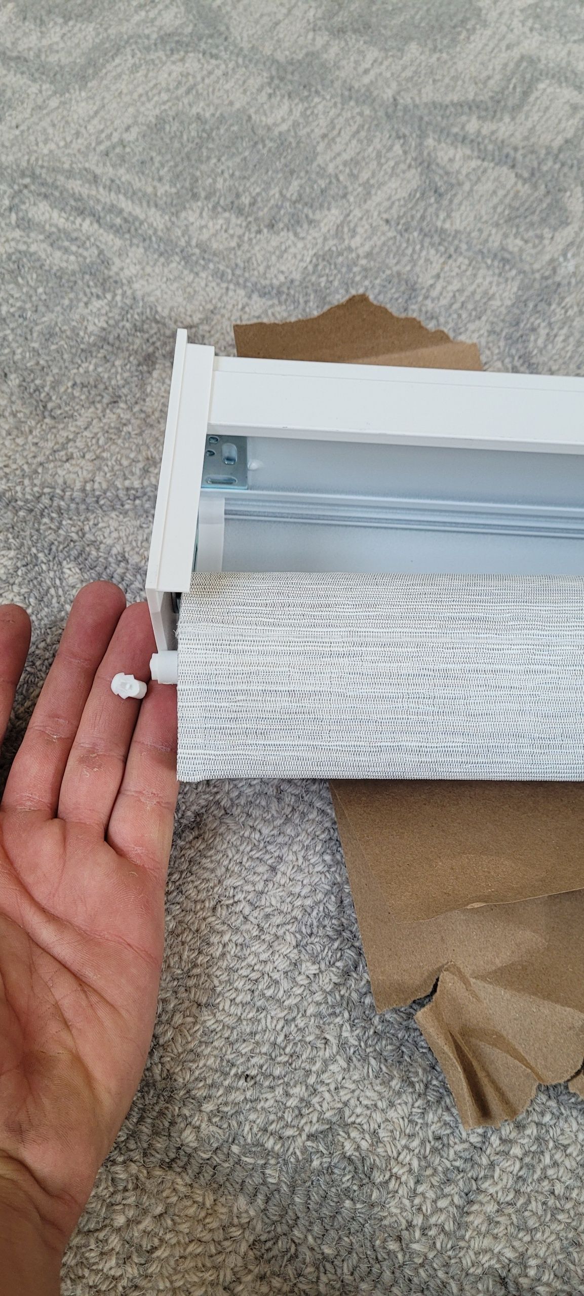 Hand holding a white roller blind mechanism; grey fabric and beige paper on a grey carpet.