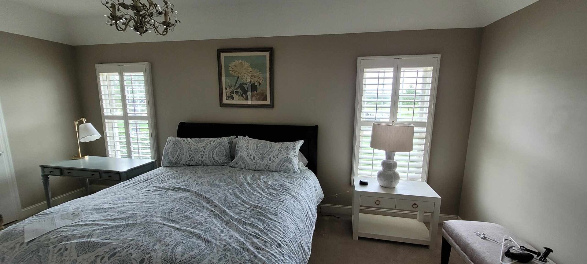 Bedroom with bed, nightstands, windows, and chandelier. Beige walls and white shutters.
