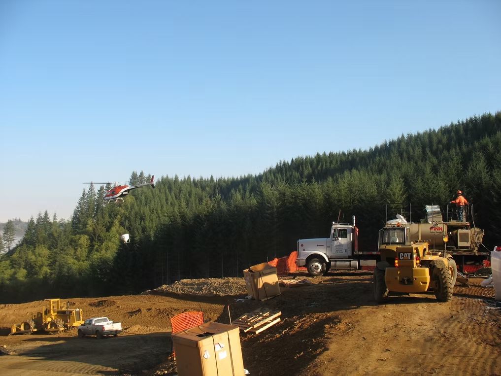 A helicopter flies over a logging site with heavy machinery and trucks parked on a dirt lot near a coniferous forest.