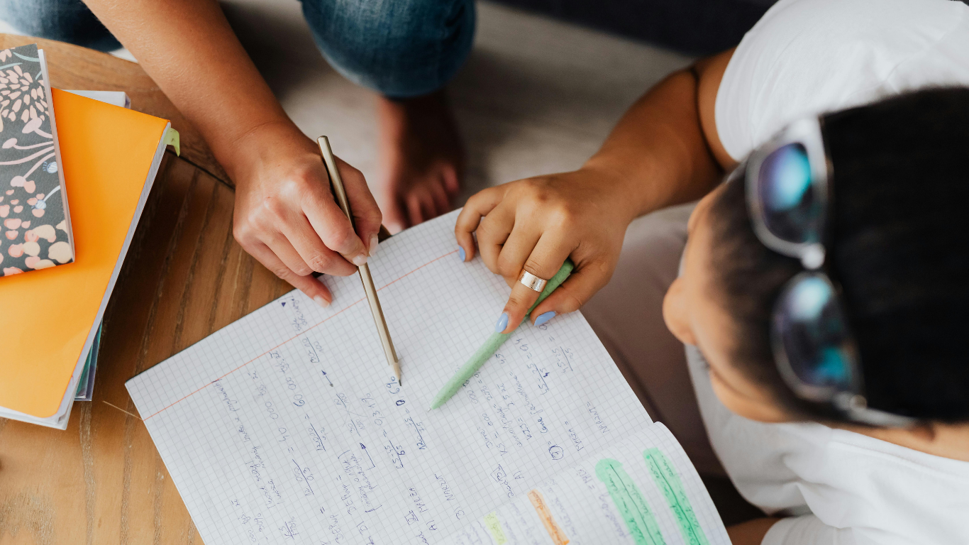 Two people study together, pointing at handwritten notes on a document resting on a wooden table beside books.