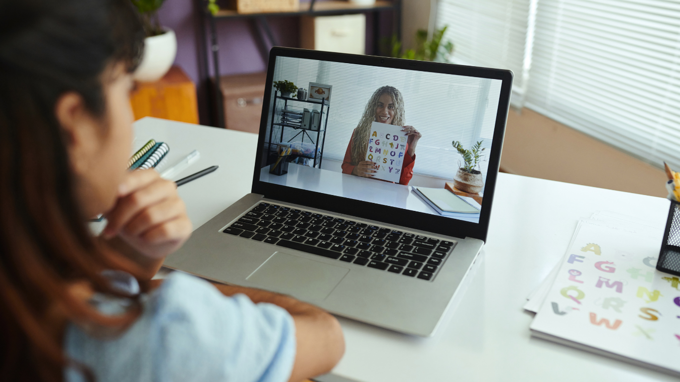 A student watches a virtual teacher holding a chart during a video lesson on a laptop.