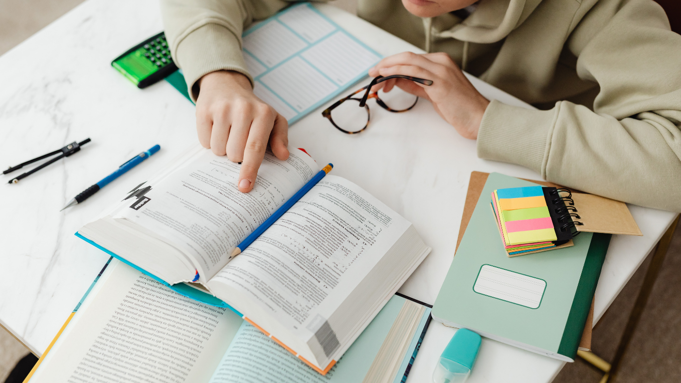 A person in a beige hoodie studies at a desk with an open textbook, glasses, pens, and a notebook.