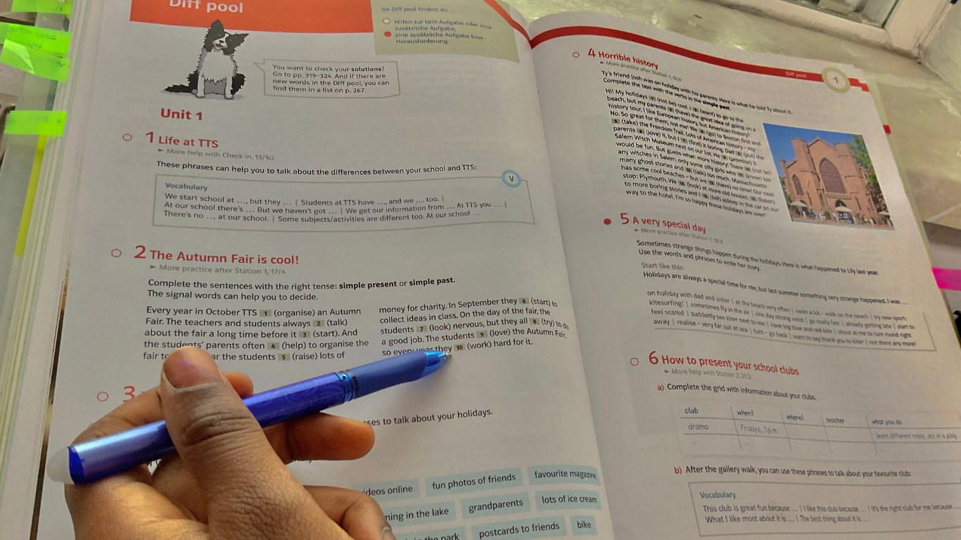 A hand holds a blue pen over an open textbook page featuring educational text and an image of a cathedral.