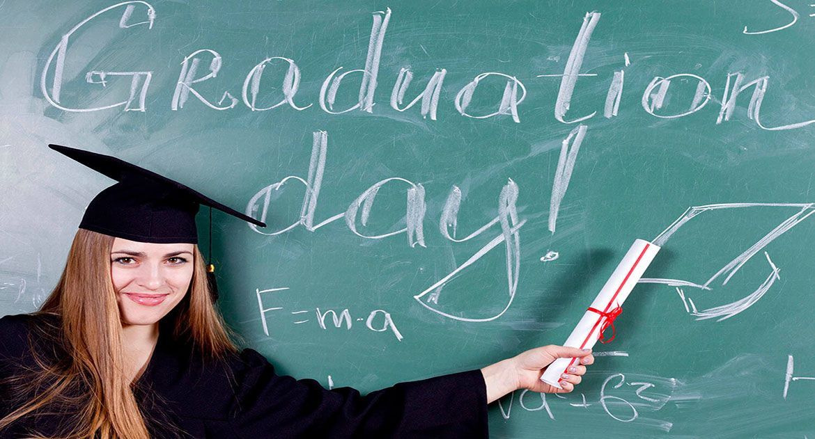 A smiling graduate in a cap and gown holds a diploma in front of a chalkboard with