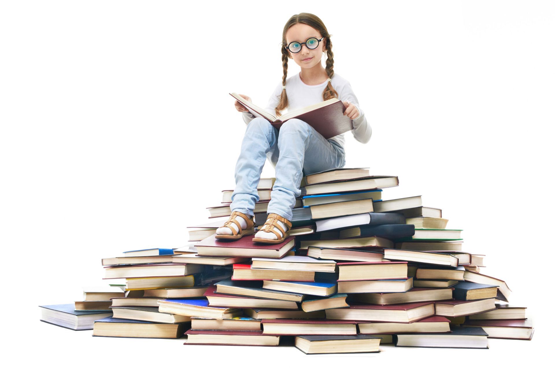A person with glasses and pigtails reads an open book while sitting atop a large, disorganized stack of various books.