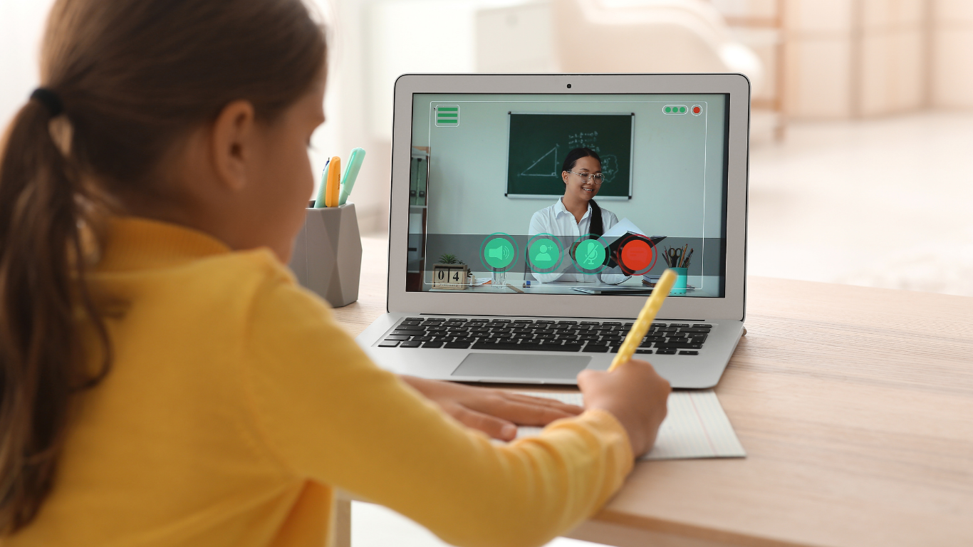 A person in a yellow shirt writes on paper while watching a teacher during a virtual lesson on a laptop.