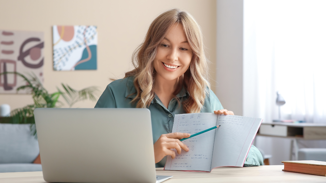 A person sitting at a desk with a laptop, smiling and pointing at an open notebook during a video call.