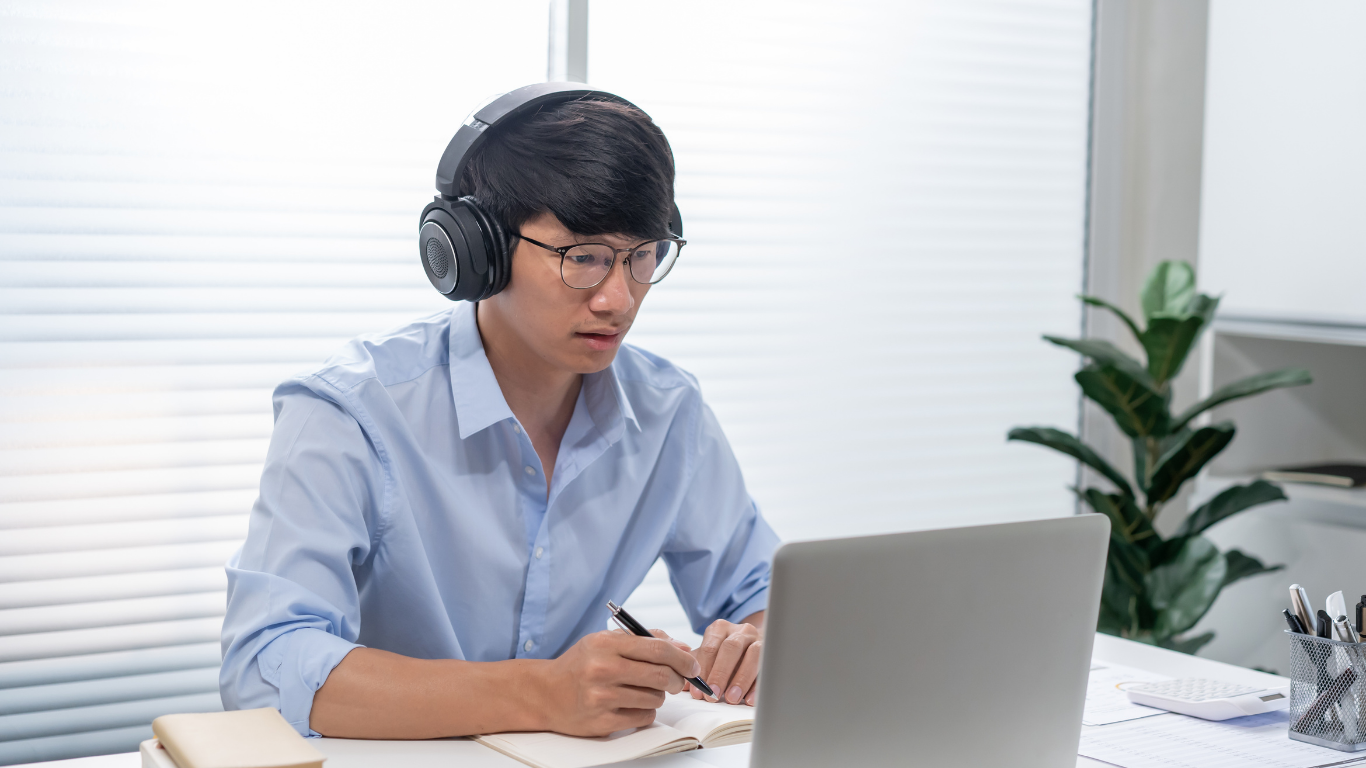 A person wearing headphones and glasses works at a desk, writing in a notebook while looking at a laptop screen.