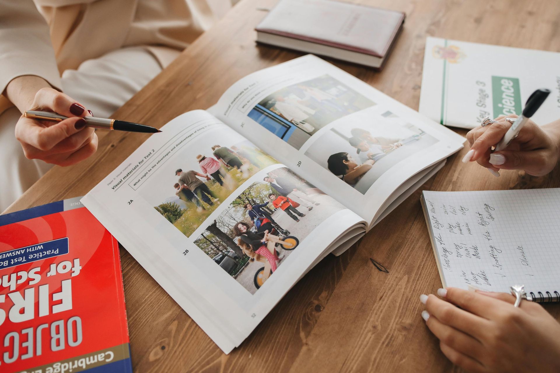 Two people study at a wooden table, pointing at a textbook with photographs while taking notes on a notepad.