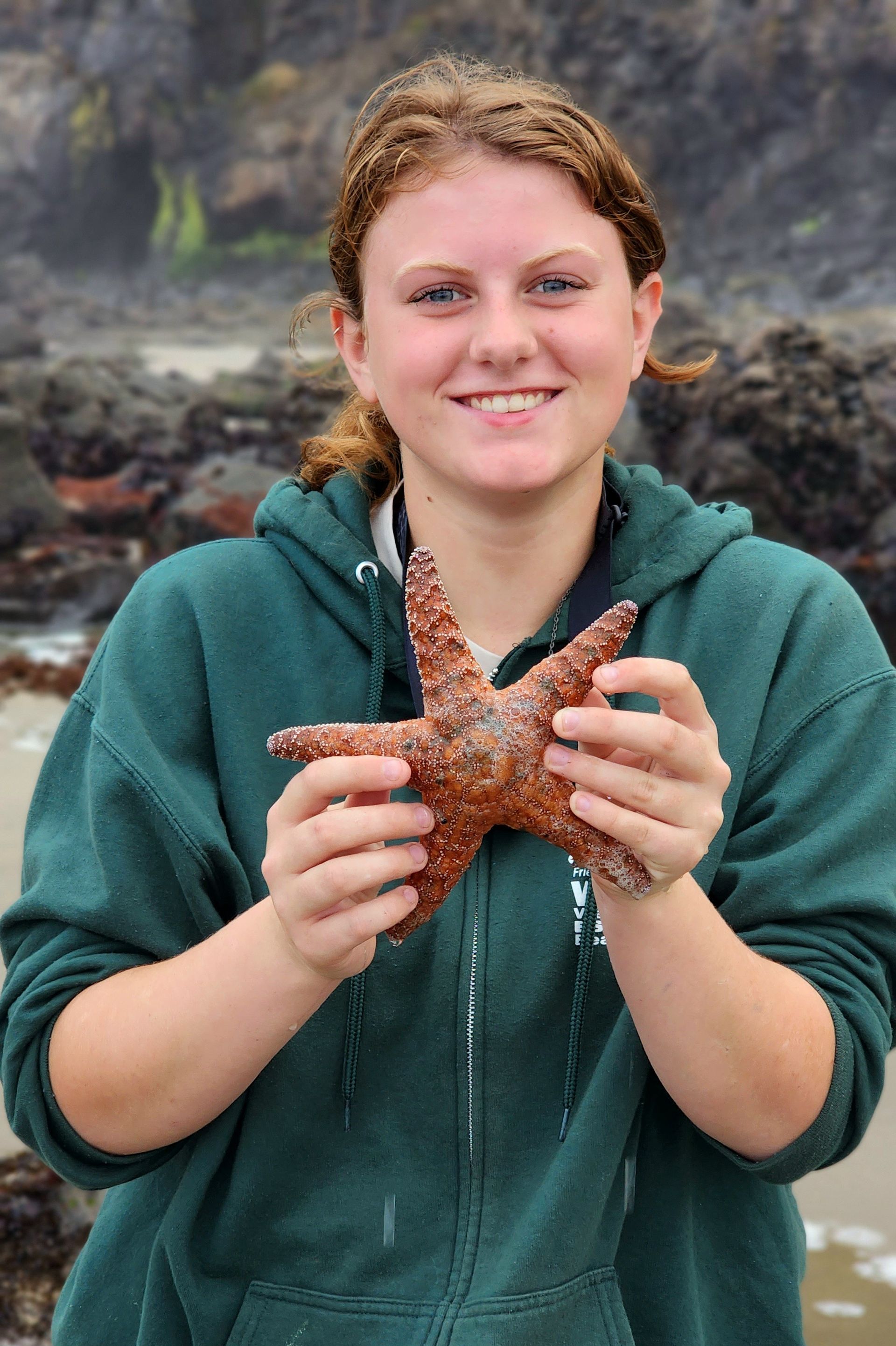 Photo of 2024 WEBS Scholarship Recipient, Naomi Travis, smiling and standing on the beach wearing a green zip-up hoodie sweatshirt while holding a orange Ochre Sea Star. 