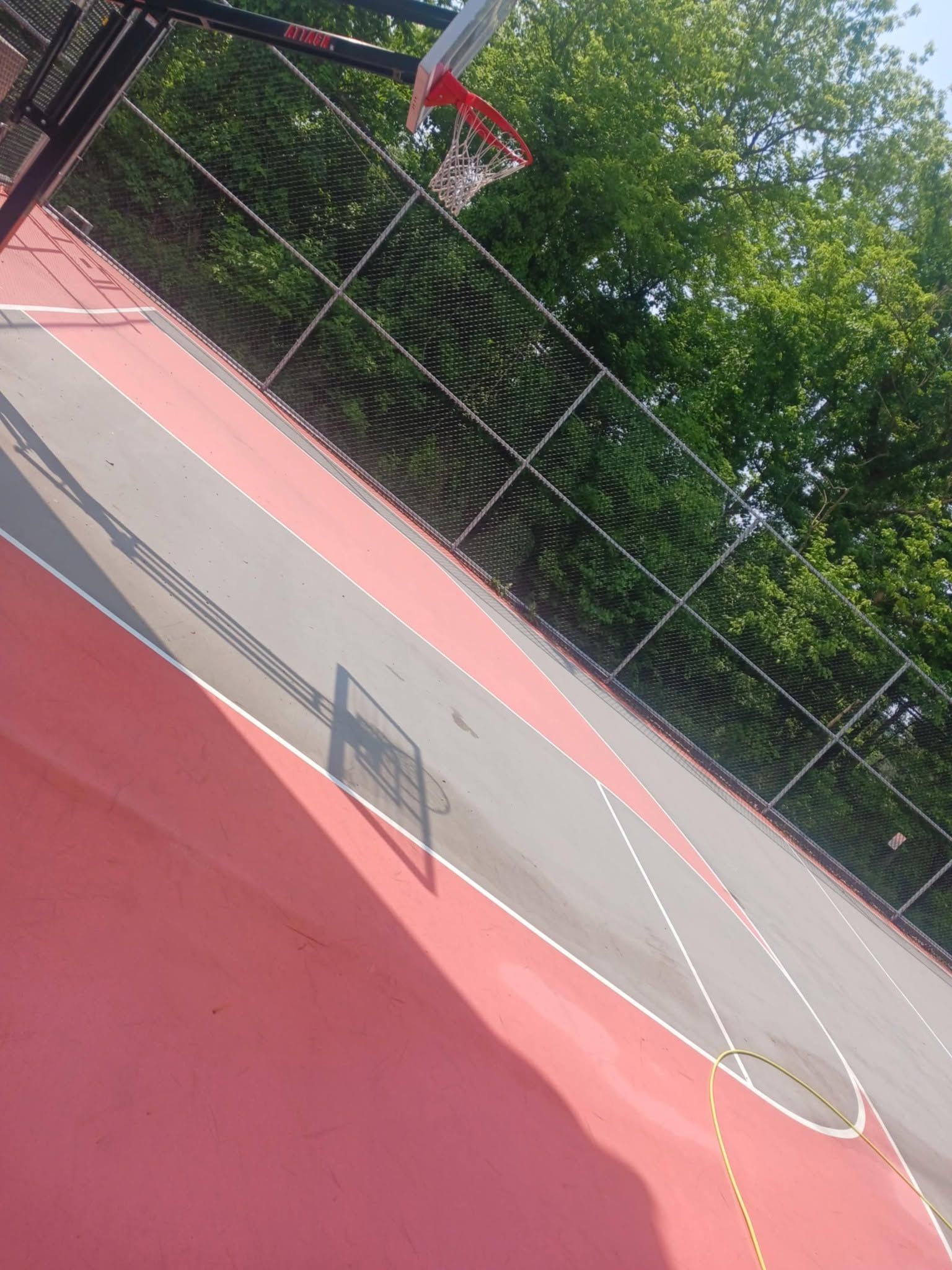 Tilted view of an empty outdoor basketball court with a hoop, red surface, and trees in the background