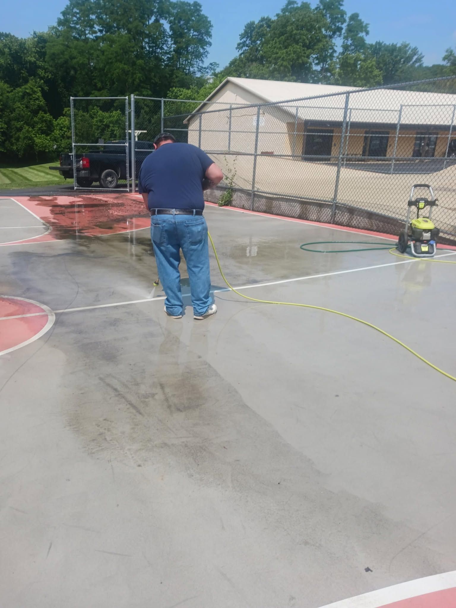 Person standing on a wet court near a fenced area and small building, with a child on a tricycle nearby.