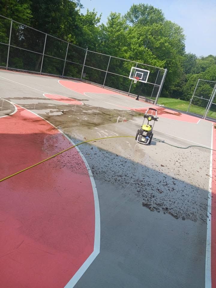 Child riding a toy car on a wet outdoor basketball court with trees and a hoop in the background