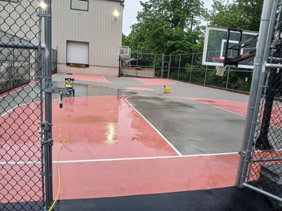 Outdoor basketball court with wet red and gray pavement beside a chain-link fence and building.