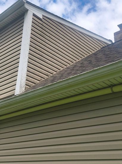 Close-up of a house roof with tan siding, green eaves, and a white gable against a partly cloudy sky