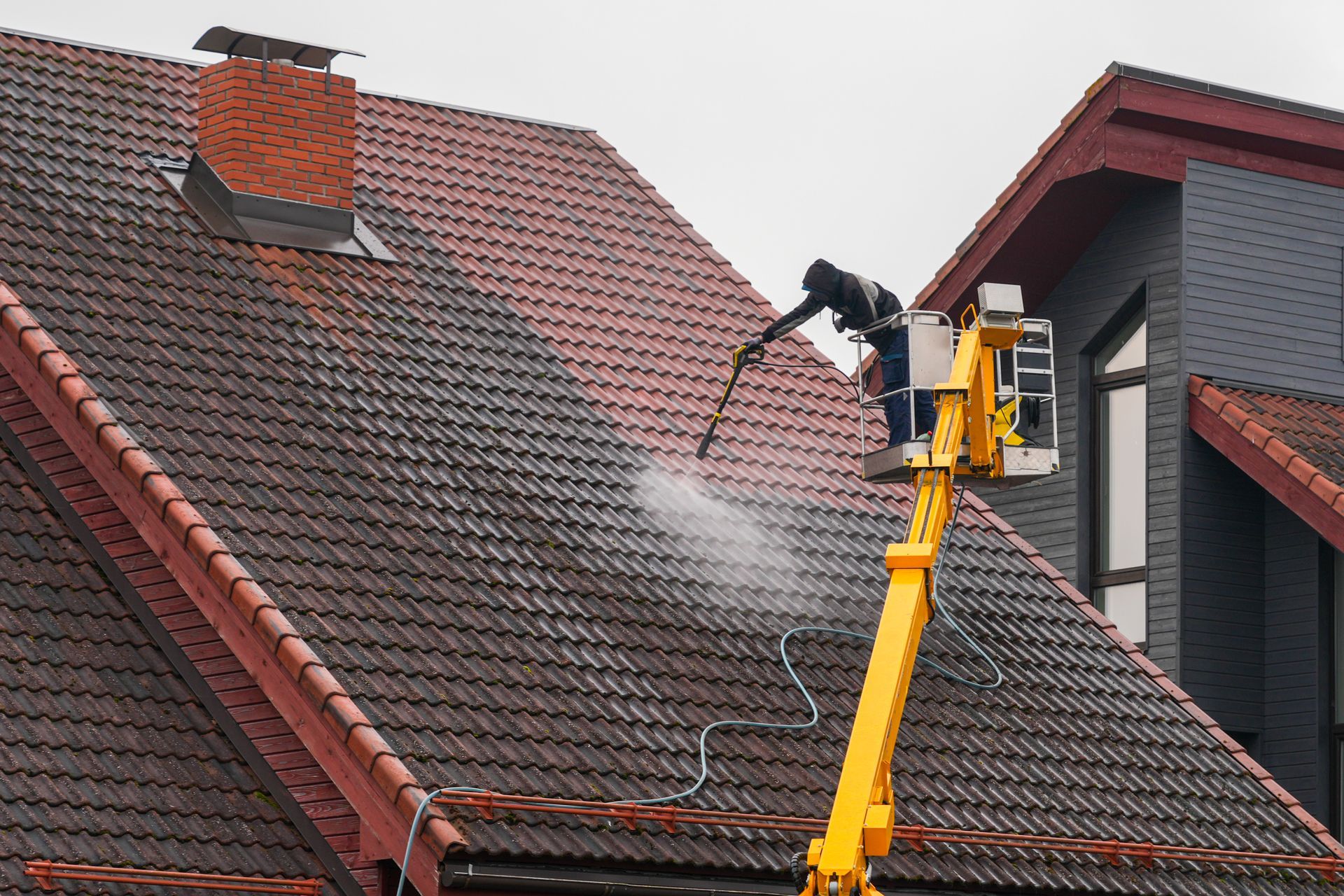 Worker power-washing a red-tiled roof from a yellow lift