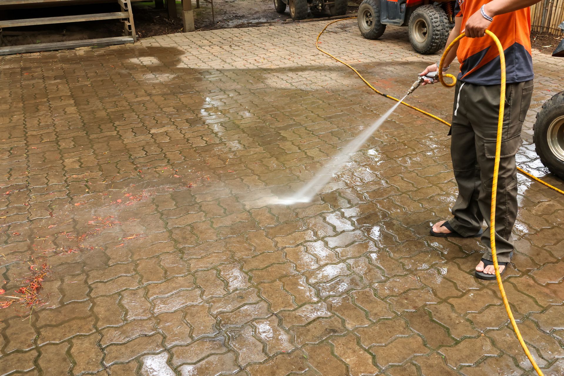 Worker pressure-washing a wet brick driveway with a hose and spray wand