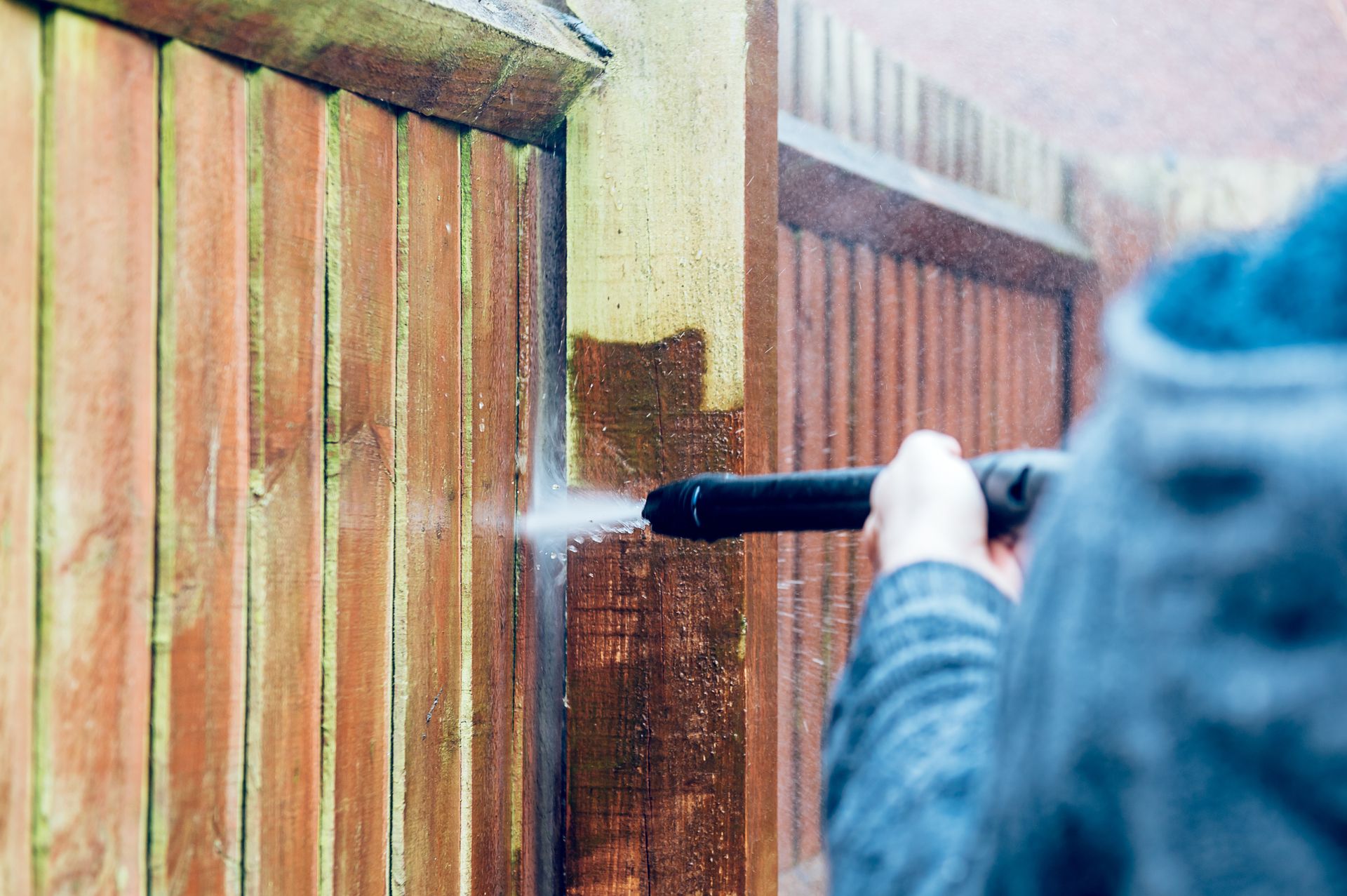 Person pressure-washing a wooden fence with a spray nozzle outdoors