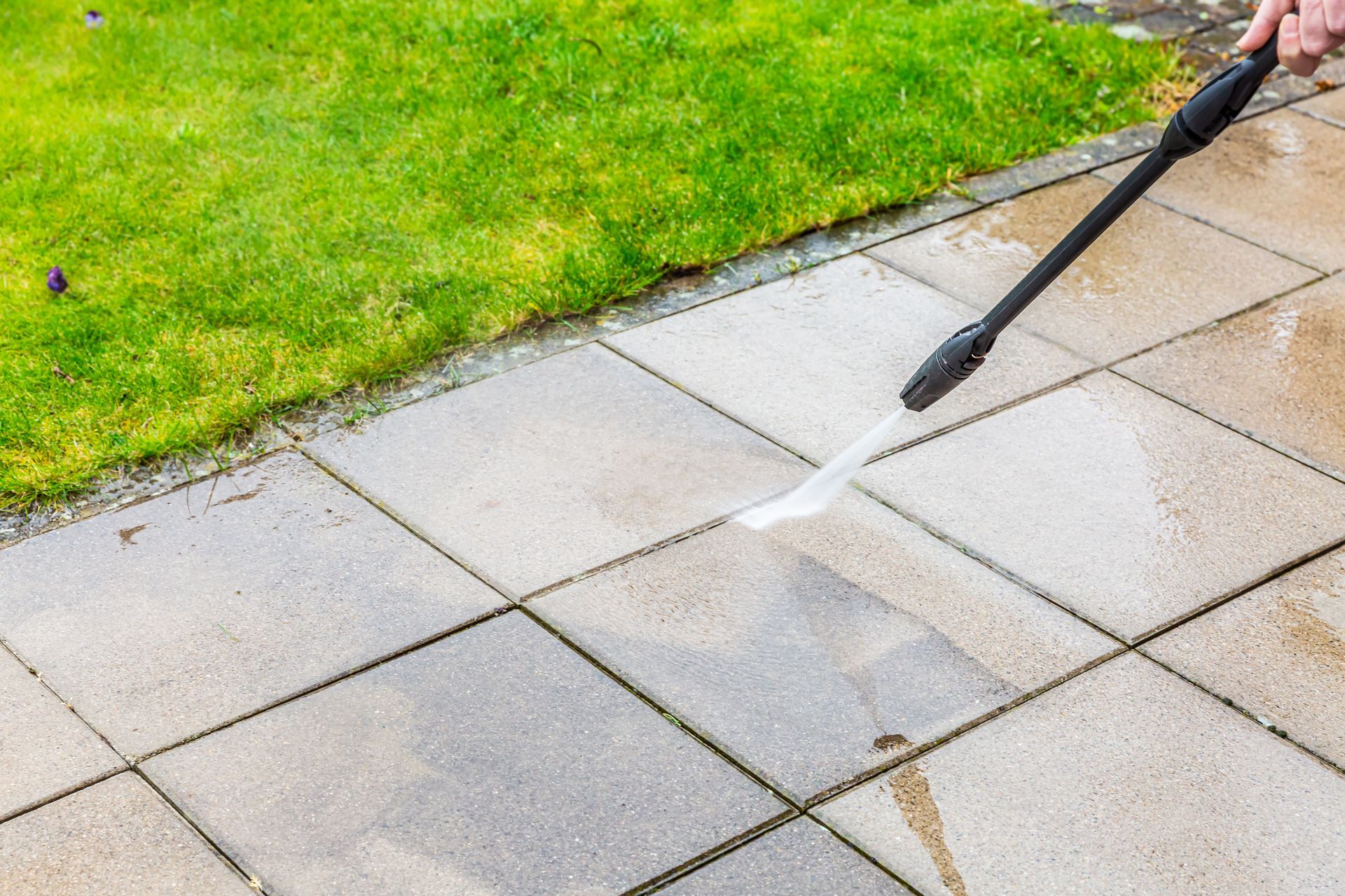 Person spraying water on a wet concrete patio beside green grass