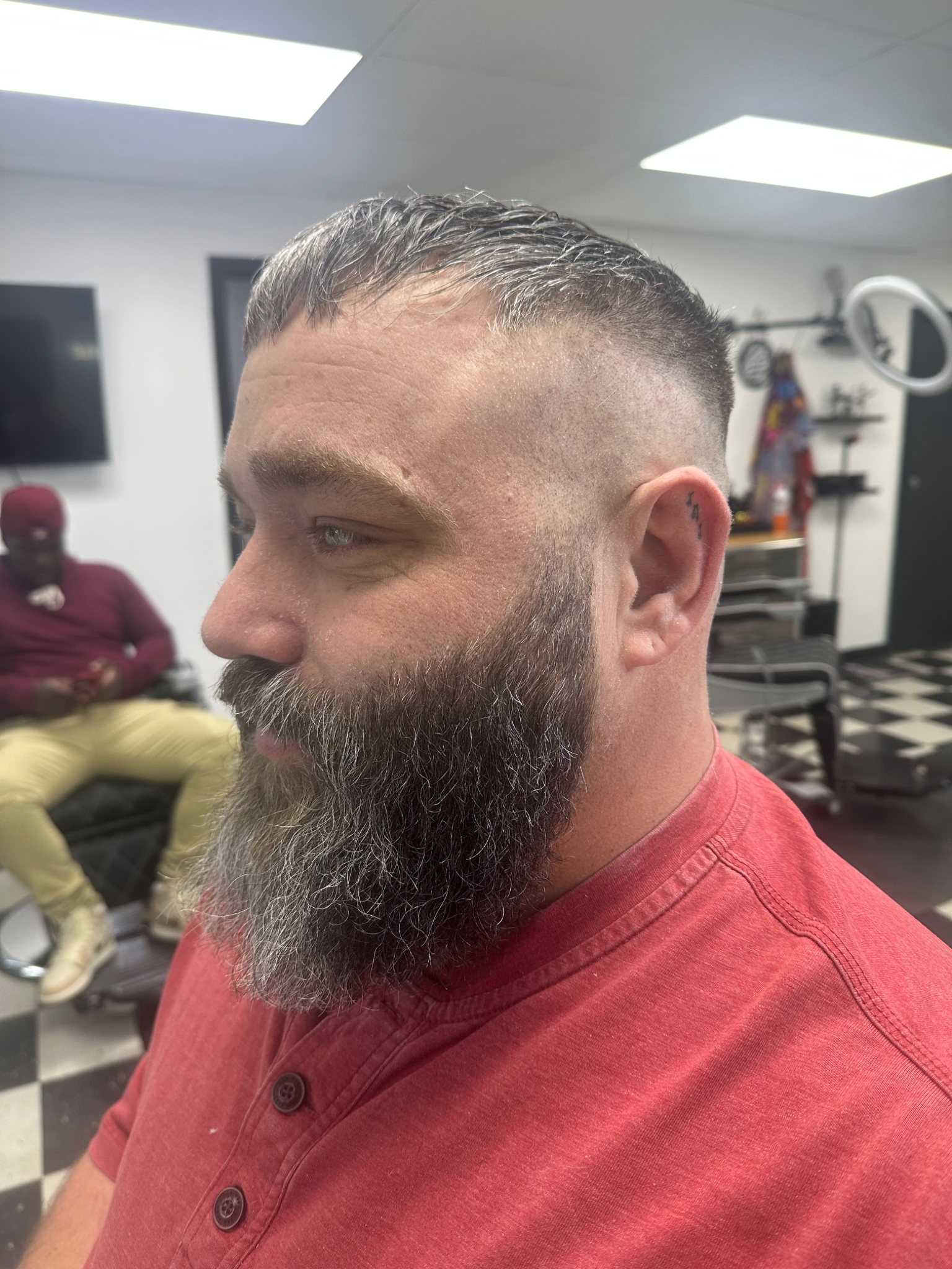 Man with a faded haircut and beard covered in hair clippings, in a barbershop.