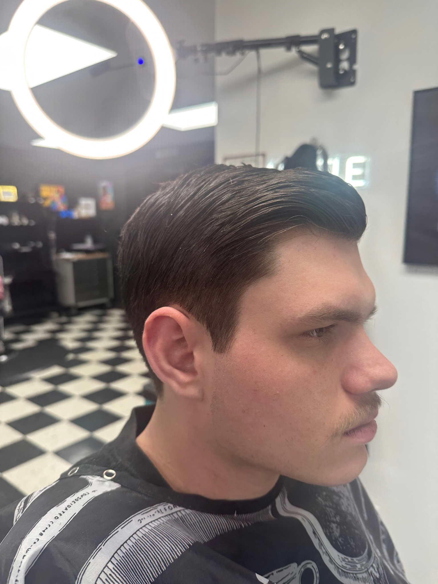 Man with slicked-back, wavy dark hair, wearing a patterned cape, looking to the side in a barbershop.
