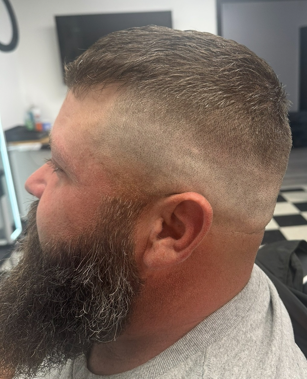 Man with faded haircut and full beard, profile view. Gray shirt, barbershop setting.