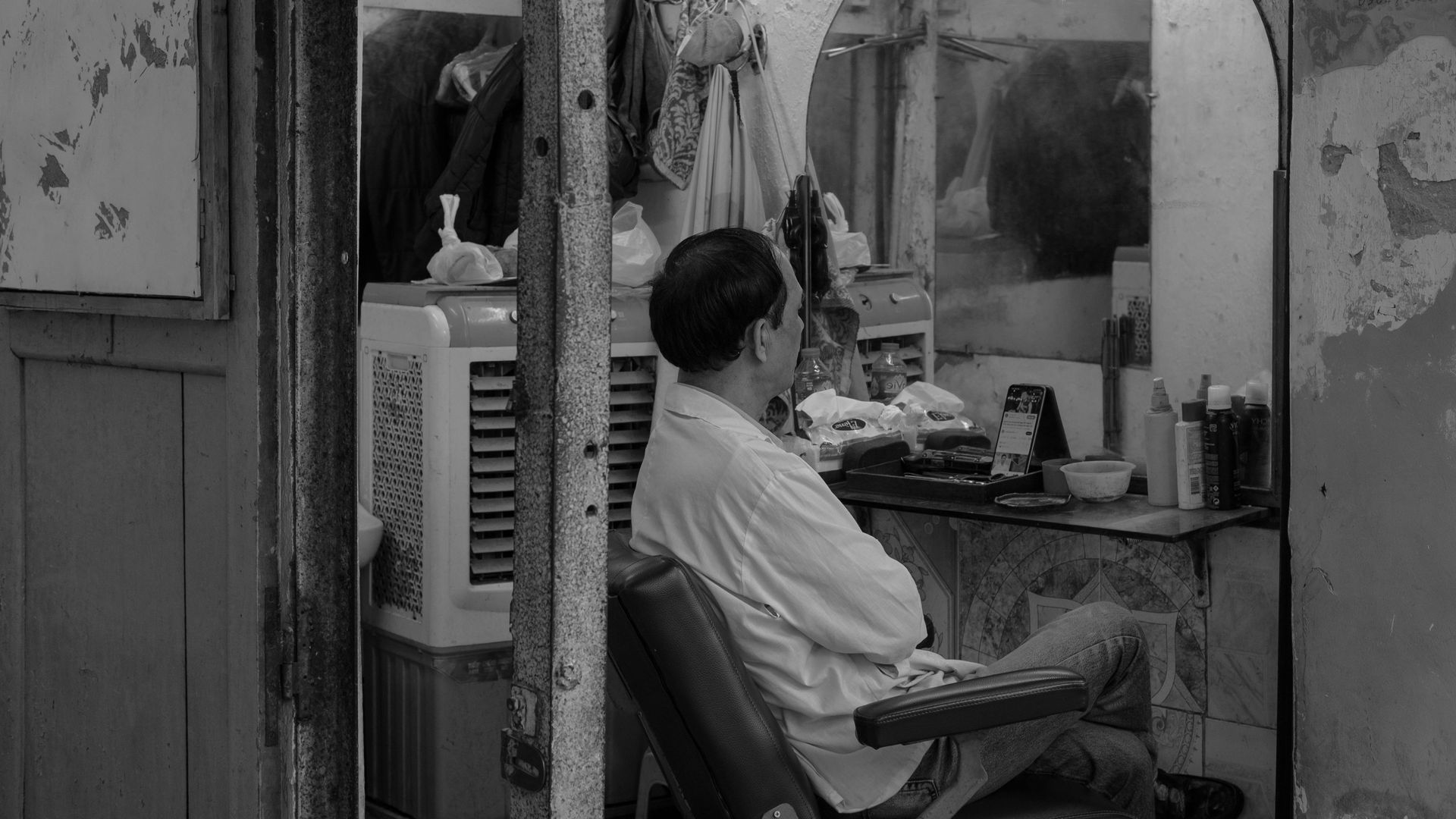 Man in white shirt sits in a dimly lit barbershop, arms crossed, looking at the mirror.