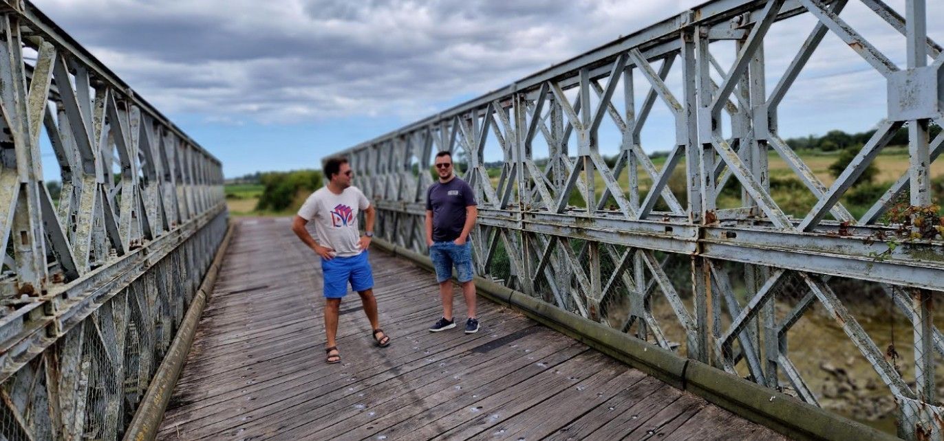 Twee mannen staan ​​op een oude metalen  Bailleybrug. Op de achtergrond is een bewolkte hemel te zien en grasvelden.