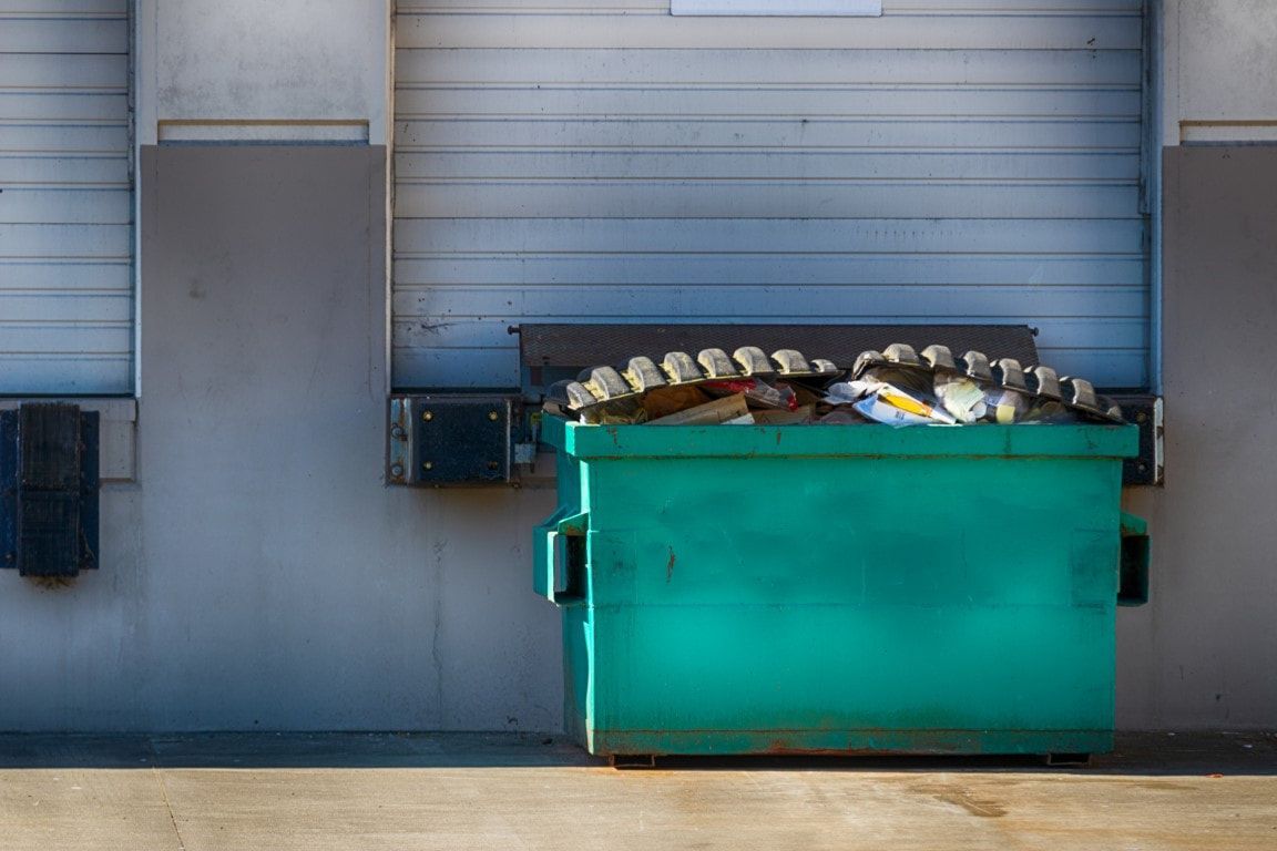 Teal dumpster overflowing with trash in front of a loading dock.
