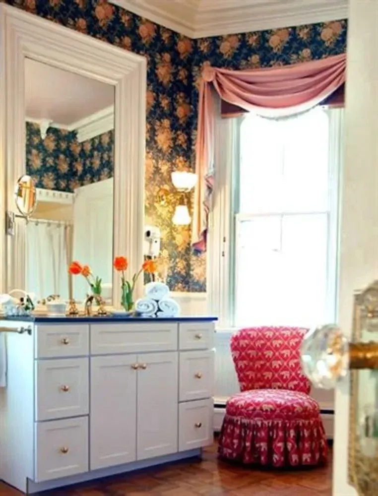 Vintage vanity with mirror, flowers, pink chair, and floral wallpaper in a bright bedroom corner