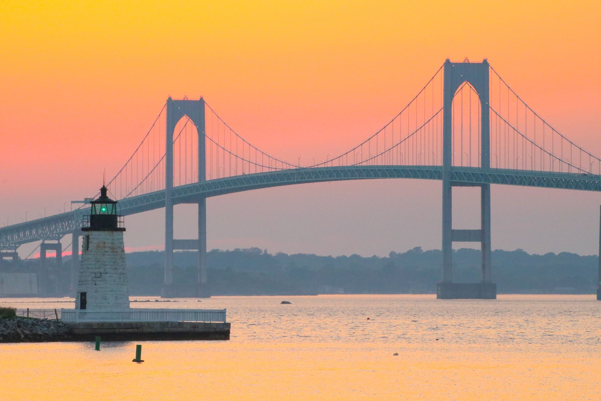 Sunset over a suspension bridge across calm water, with a small lighthouse in the foreground.