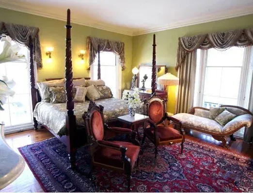 Elegant bedroom with four-poster bed, antique chairs, patterned rugs, and large windows with draped curtains