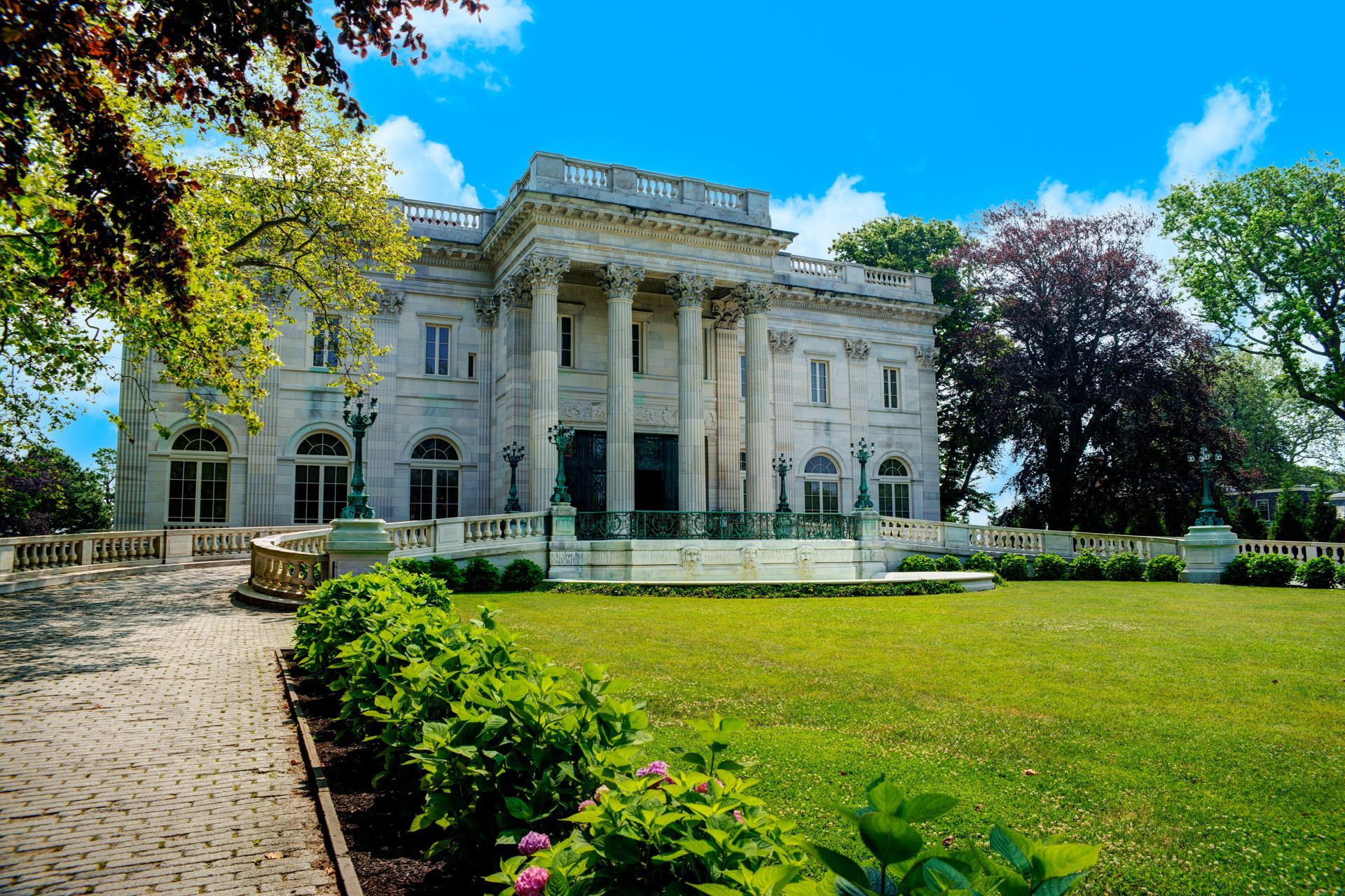 Grand white neoclassical mansion with columns, green lawn, and trees under a blue sky
