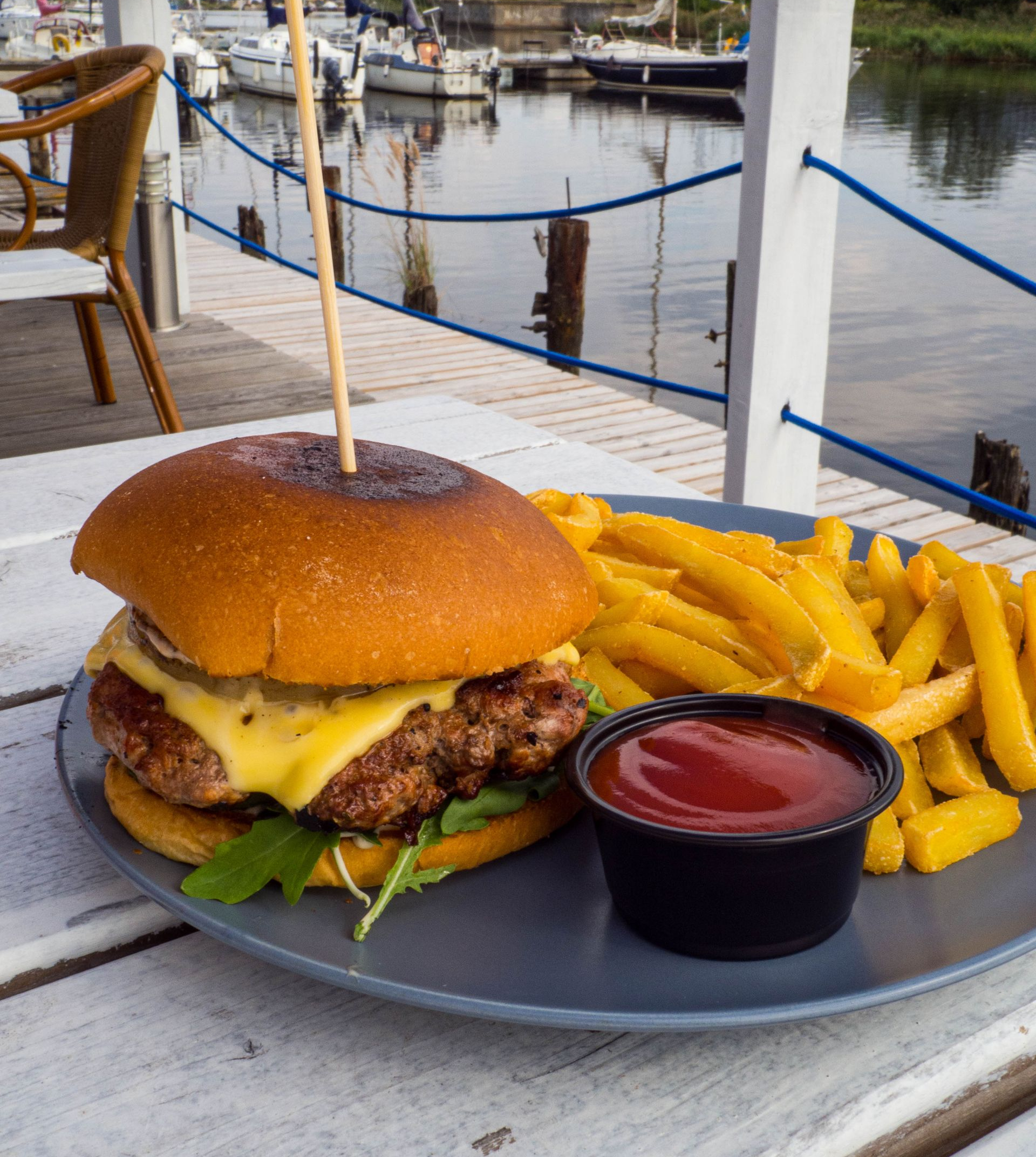 Cheeseburger, fries, and ketchup on a plate by the water at an outdoor café