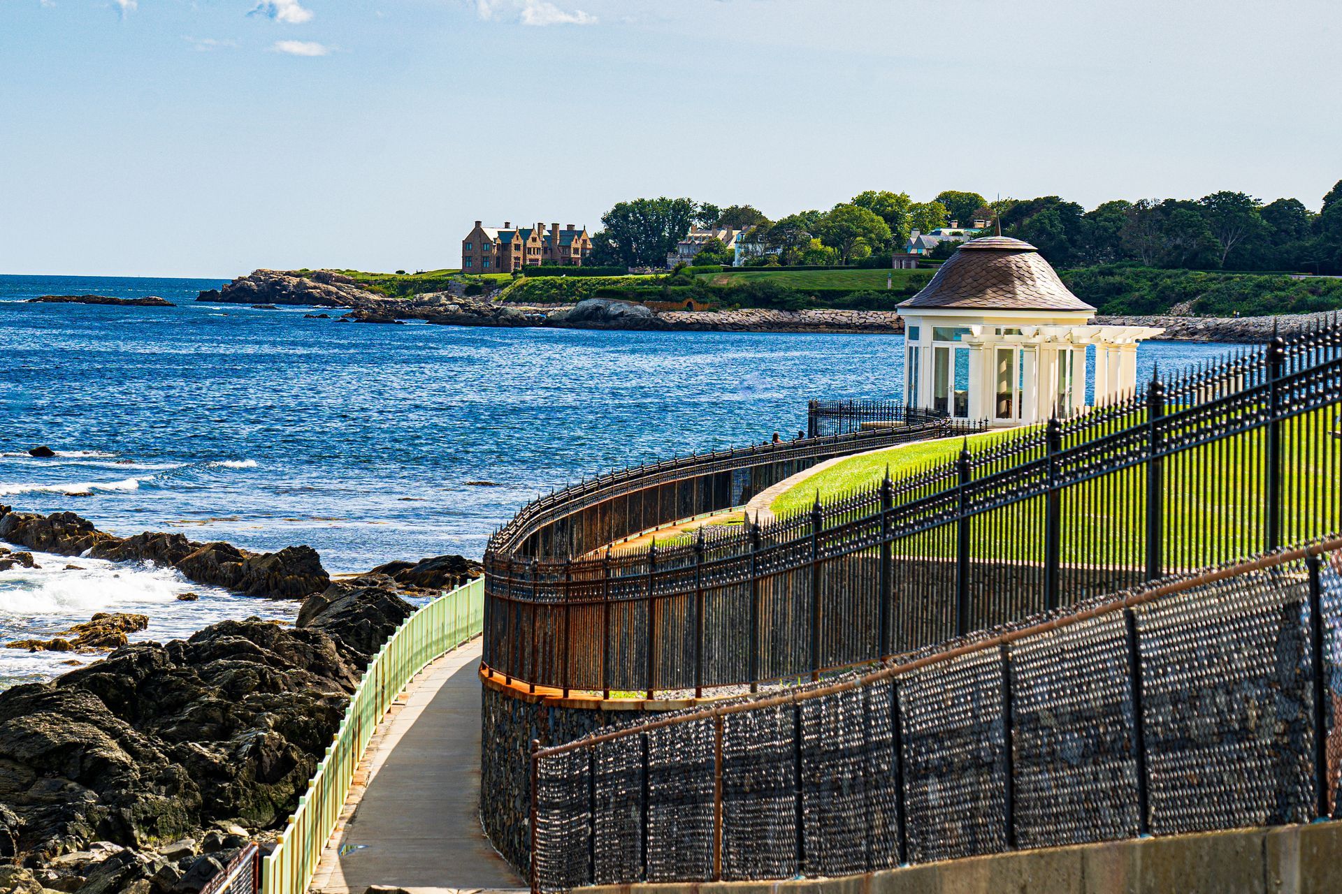 Coastal walkway beside blue water, with a black fence and a small pavilion on a rocky shore.