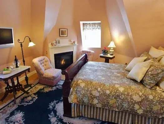 Cozy bedroom with floral bedspread, fireplace, armchair, bedside lamps, and a window with curtains.