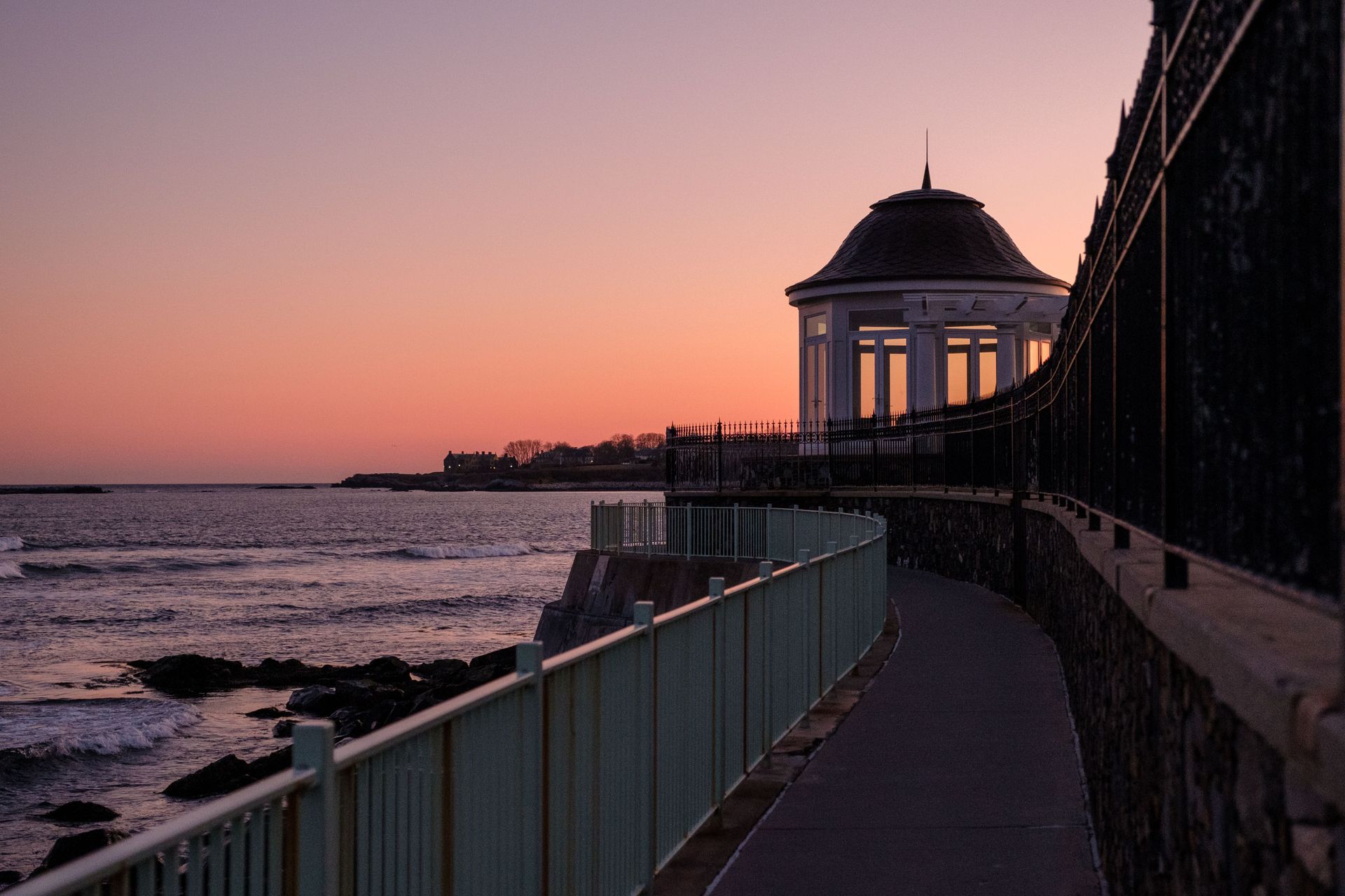 Sunset over a seaside walkway with a gazebo, railing, and calm ocean beside a stone wall