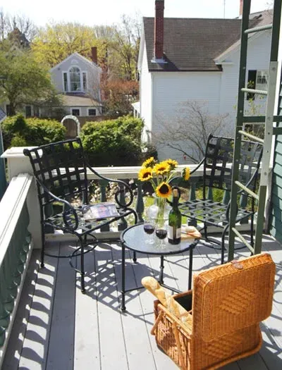 Sunlit patio with black metal table and chairs, sunflowers, and a wicker chair overlooking a garden and houses